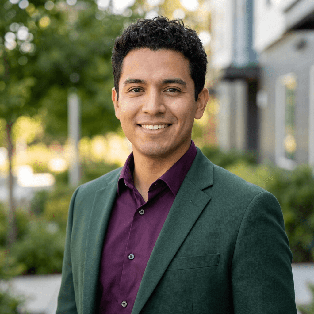 Smiling man with dark curly hair wearing a green blazer and purple button-down shirt.